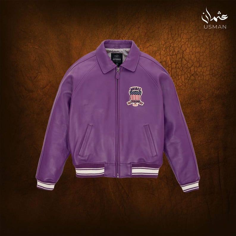 May include: A purple leather jacket with a bomber style collar and a zipper closure. The jacket features a patch with a patriotic design on the chest and white and black striped cuffs and hem. The jacket is displayed on a brown background.