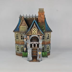 Op de afbeelding: Een gedetailleerd miniatuurhuis met een bruin, blauw en beige kleurenschema. Het huis heeft een stenen gevel, een gewelfde deuropening en een schoorsteen. De ramen hebben een gele gloed en er zijn decoratieve lantaarns.