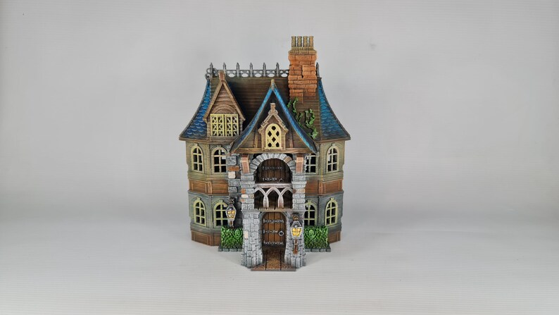 Op de afbeelding: Een gedetailleerd miniatuurhuis met een bruin, blauw en beige kleurenschema. Het huis heeft een stenen gevel, een gewelfde deuropening en een schoorsteen. De ramen hebben een gele gloed en er zijn decoratieve lantaarns.