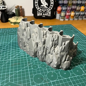 Puede incluir: Una pieza de terreno gris impresa en 3D que representa un acantilado rocoso con un dise&ntilde;o curvo y aberturas arqueadas. Una funda negra con el texto "WAR RAVEN" y un emblema blanco est&aacute; en el fondo. La pieza de terreno est&aacute; sobre una esterilla de corte verde.