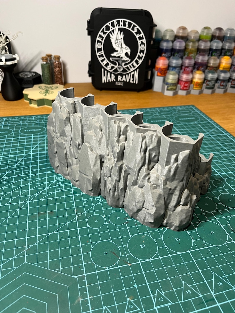 Puede incluir: Una pieza de terreno gris impresa en 3D que representa un acantilado rocoso con un dise&ntilde;o curvo y aberturas arqueadas. Una funda negra con el texto "WAR RAVEN" y un emblema blanco est&aacute; en el fondo. La pieza de terreno est&aacute; sobre una esterilla de corte verde.