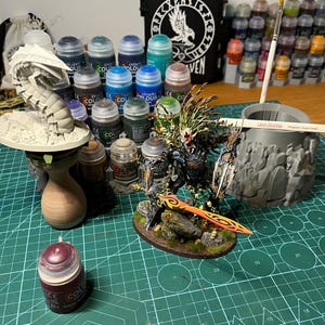 Puede incluir: Una colecci&oacute;n de suministros de pintura en miniatura, que incluye varios botes de pintura Citadel Colour, pinceles y una figura en miniatura. Se ve una funda negra con un logotipo de cuervo blanco. La escena est&aacute; sobre una esterilla de corte verde.