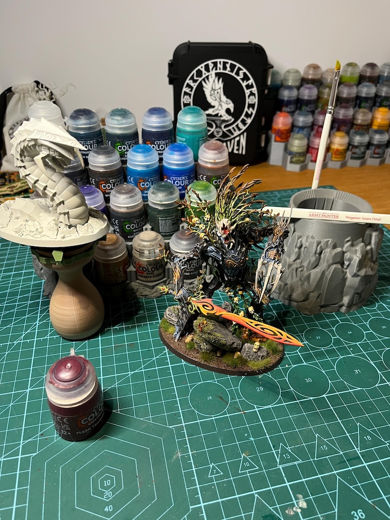 Puede incluir: Una colecci&oacute;n de suministros de pintura en miniatura, que incluye varios botes de pintura Citadel Colour, pinceles y una figura en miniatura. Se ve una funda negra con un logotipo de cuervo blanco. La escena est&aacute; sobre una esterilla de corte verde.