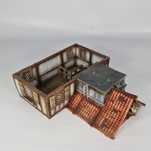 May include: A detailed miniature building model, featuring a timber-framed structure with a red-tiled roof. The model has multiple sections, including a main building and an attached roofed area. The building has windows and is designed for tabletop gaming or model enthusiasts.