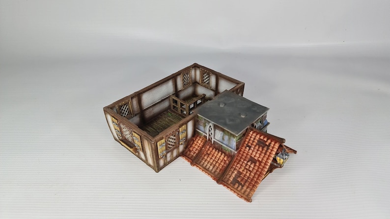 May include: A detailed miniature building model, featuring a timber-framed structure with a red-tiled roof. The model has multiple sections, including a main building and an attached roofed area. The building has windows and is designed for tabletop gaming or model enthusiasts.