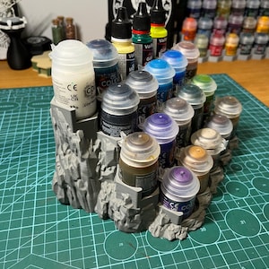 Puede incluir: Un organizador de botes de pintura gris impreso en 3D que contiene varios botes de pintura. Los botes est&aacute;n llenos de diferentes colores, incluyendo amarillo, azul y marr&oacute;n. Una funda negra con texto blanco est&aacute; en el fondo. El organizador est&aacute; sobre una esterilla de corte verde.