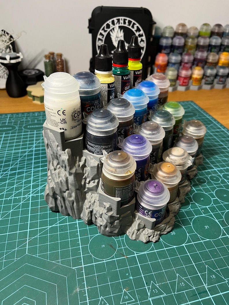 Puede incluir: Un organizador de botes de pintura gris impreso en 3D que contiene varios botes de pintura. Los botes est&aacute;n llenos de diferentes colores, incluyendo amarillo, azul y marr&oacute;n. Una funda negra con texto blanco est&aacute; en el fondo. El organizador est&aacute; sobre una esterilla de corte verde.