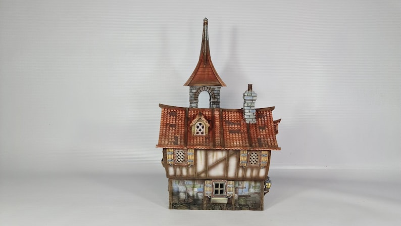 May include: A detailed model of a medieval-style building with a red-tiled roof, a tall steeple, and a chimney. The walls are a combination of light-coloured wood and dark brown beams, with small windows and decorative elements. The model is approximately 20 cm tall.