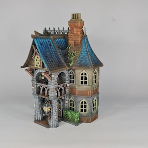 Op de afbeelding: Een gedetailleerd miniatuur fantasiehuis met een blauw pannendak, gewelfde ramen en een stenen gevel. Het huis heeft een schoorsteen en decoratieve afwerking, met een kleine groene struik aan de basis. Het kleurenpalet omvat tinten blauw, bruin en grijs.