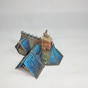 Op de afbeelding: Miniatuurmodel van een huis met een blauw pannendak en een bakstenen schoorsteen. Het dak heeft een gedetailleerde textuur en de schoorsteen heeft een verweerde uitstraling. Het huis heeft een klein raam en een decoratief hek.