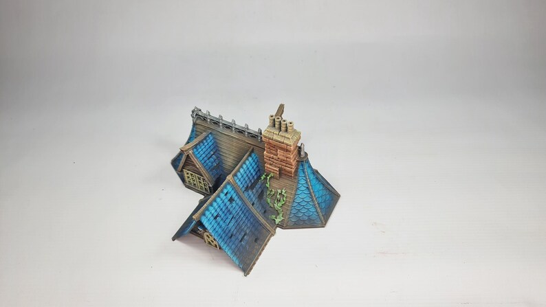 Op de afbeelding: Miniatuurmodel van een huis met een blauw pannendak en een bakstenen schoorsteen. Het dak heeft een gedetailleerde textuur en de schoorsteen heeft een verweerde uitstraling. Het huis heeft een klein raam en een decoratief hek.