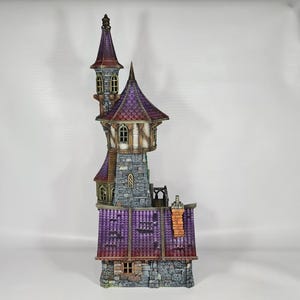 以下が含まれることがあります： 高い塔と複数の小塔を備えた、詳細なミニチュアファンタジー城。城は石の基部、紫色の屋根、茶色とタンのアクセントが特徴です。塔には小さな窓と尖った上部があります。