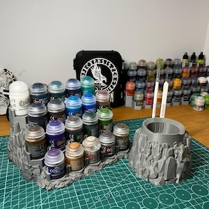 Puede incluir: Un organizador de pintura gris impreso en 3D con un dise&ntilde;o de terreno rocoso. Contiene varios botes de pintura y dos pinceles. Una funda negra con un emblema blanco est&aacute; en el fondo, junto con m&aacute;s botes de pintura.