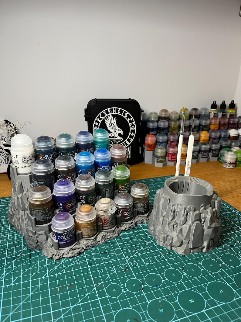 Puede incluir: Un organizador de pintura gris impreso en 3D con un dise&ntilde;o de terreno rocoso. Contiene varios botes de pintura y dos pinceles. Una funda negra con un emblema blanco est&aacute; en el fondo, junto con m&aacute;s botes de pintura.