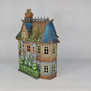 Op de afbeelding: Een gedetailleerd miniatuurhuis met een verweerde uitstraling. Het model heeft een bruin dak met blauwe accenten, stenen muren en meerdere ramen. Groene bladeren voegen een vleugje natuur toe. De schoorsteen is roodbruin.