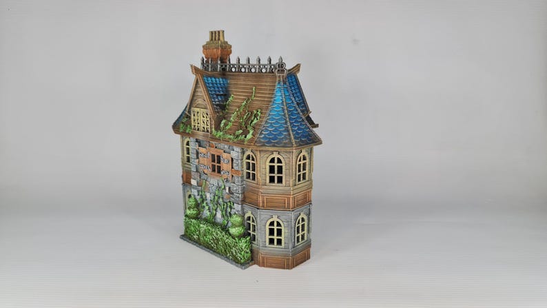 Op de afbeelding: Een gedetailleerd miniatuurhuis met een verweerde uitstraling. Het model heeft een bruin dak met blauwe accenten, stenen muren en meerdere ramen. Groene bladeren voegen een vleugje natuur toe. De schoorsteen is roodbruin.