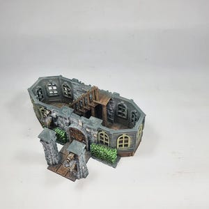 Op de afbeelding: Een miniatuur kasteelmodel met grijze stenen muren, boogvensters en een houten trap. Het model heeft een gedetailleerd exterieur met een poort, pilaren en groene struiken, ideaal voor tabletop gaming of display.