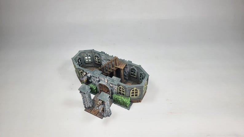 Op de afbeelding: Een miniatuur kasteelmodel met grijze stenen muren, boogvensters en een houten trap. Het model heeft een gedetailleerd exterieur met een poort, pilaren en groene struiken, ideaal voor tabletop gaming of display.