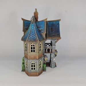 Op de afbeelding: Een gedetailleerd miniatuurhuis met een blauw en bruin dak, gewelfde ramen en decoratieve lantaarns. Het huis heeft een stenen toren en een grillig ontwerp, perfect voor verzamelaars of als decoratief stuk.