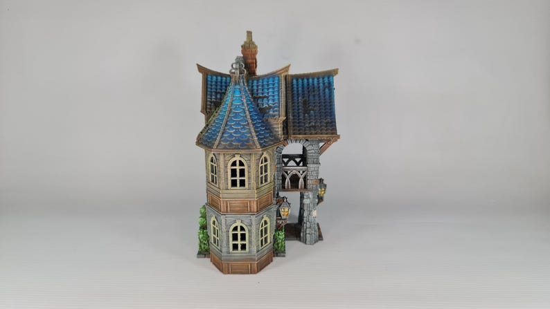 Op de afbeelding: Een gedetailleerd miniatuurhuis met een blauw en bruin dak, gewelfde ramen en decoratieve lantaarns. Het huis heeft een stenen toren en een grillig ontwerp, perfect voor verzamelaars of als decoratief stuk.