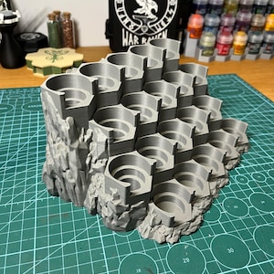 Puede incluir: Un soporte gris para botes de pintura impreso en 3D con m&uacute;ltiples ranuras circulares, dise&ntilde;ado para parecerse a una estructura rocosa. El soporte est&aacute; sobre una esterilla de corte verde, con una funda negra en el fondo que dice "WAR RAVEN".