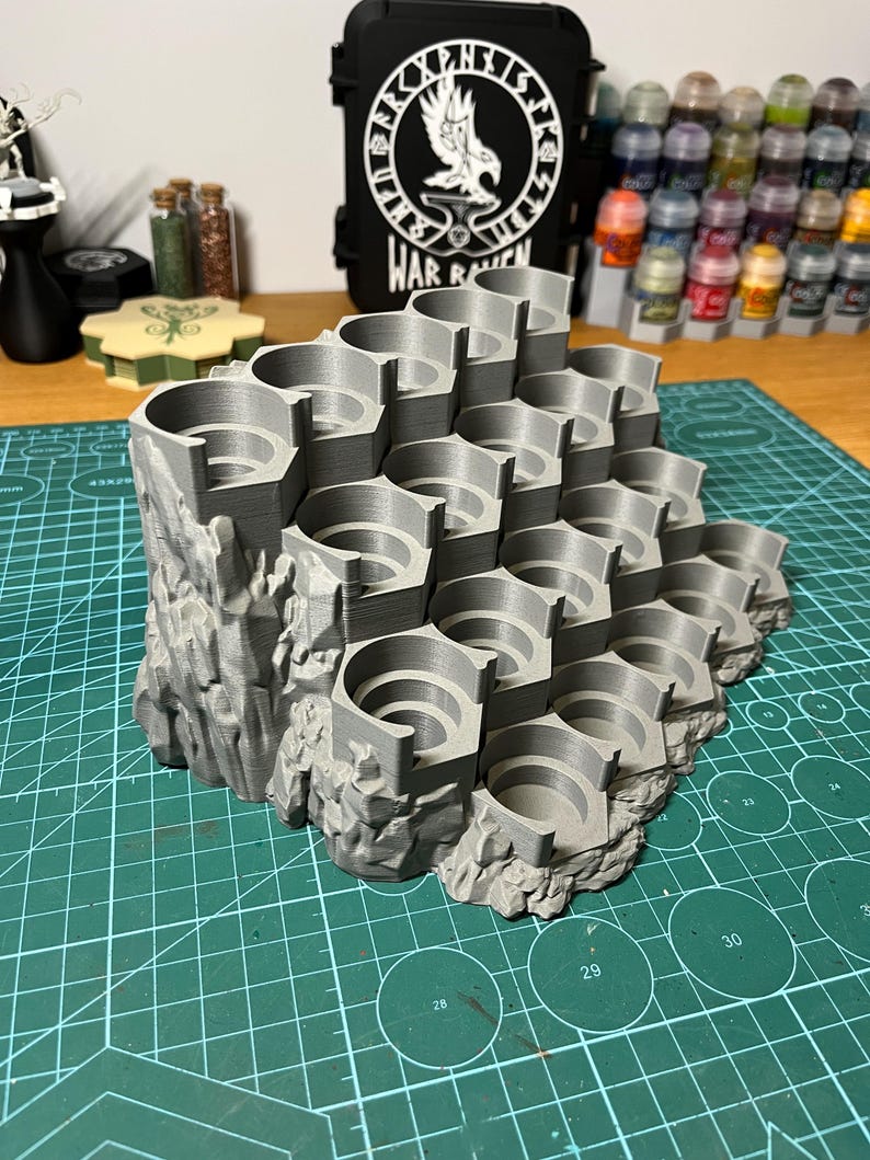 Puede incluir: Un soporte gris para botes de pintura impreso en 3D con m&uacute;ltiples ranuras circulares, dise&ntilde;ado para parecerse a una estructura rocosa. El soporte est&aacute; sobre una esterilla de corte verde, con una funda negra en el fondo que dice "WAR RAVEN".