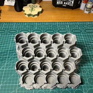 Puede incluir: Un soporte gris para botes de pintura, impreso en 3D, con m&uacute;ltiples compartimentos hexagonales. El soporte est&aacute; dise&ntilde;ado para sujetar de forma segura botes de pintura peque&ntilde;os, manteni&eacute;ndolos organizados. El soporte est&aacute; sobre una esterilla de corte verde. Otros art&iacute;culos son visibles en el fondo.