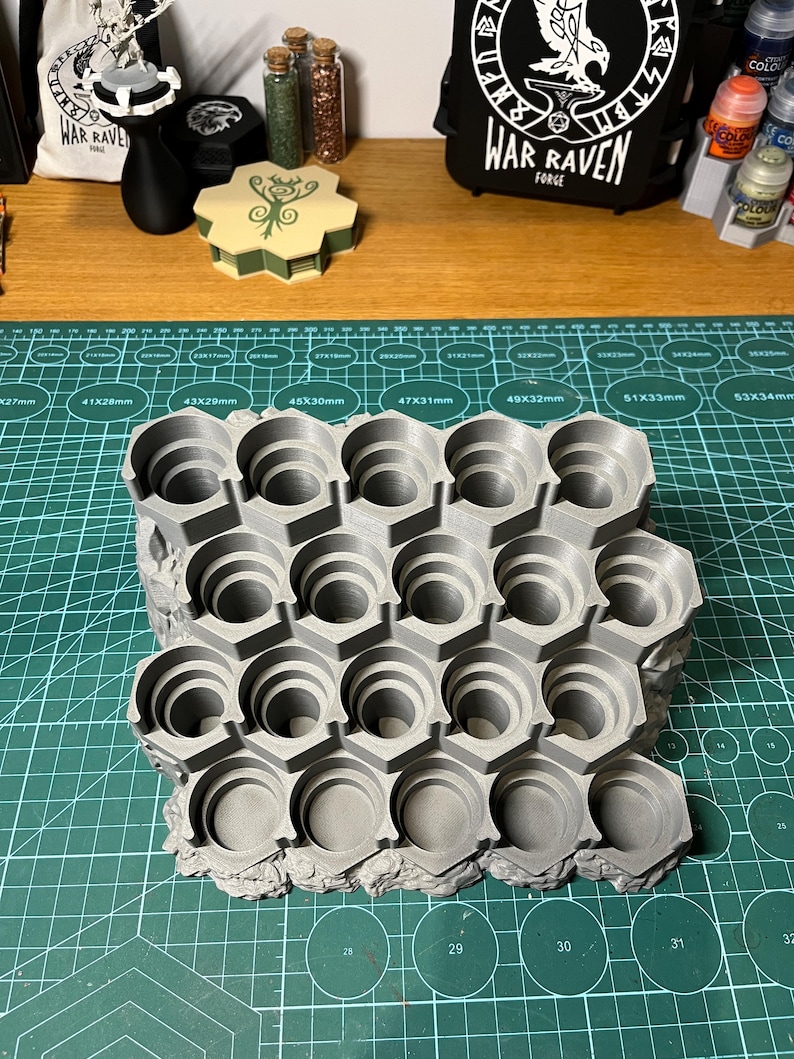 Puede incluir: Un soporte gris para botes de pintura, impreso en 3D, con m&uacute;ltiples compartimentos hexagonales. El soporte est&aacute; dise&ntilde;ado para sujetar de forma segura botes de pintura peque&ntilde;os, manteni&eacute;ndolos organizados. El soporte est&aacute; sobre una esterilla de corte verde. Otros art&iacute;culos son visibles en el fondo.