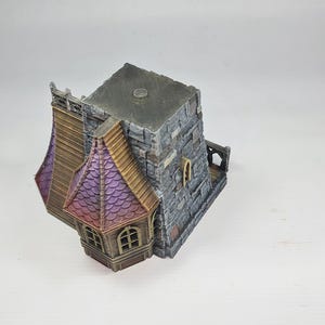 以下が含まれることがあります： グレーの石の外観、紫と茶色の屋根、小さなアーチ型の窓を備えたミニチュアファンタジービル。 このモデルは、中世またはゴシック建築様式を示唆する詳細なデザインです。 建物は白い背景に設定されています。