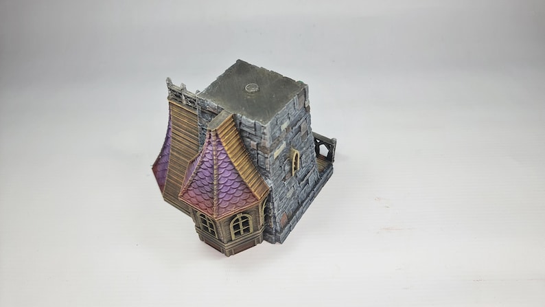 以下が含まれることがあります： グレーの石の外観、紫と茶色の屋根、小さなアーチ型の窓を備えたミニチュアファンタジービル。 このモデルは、中世またはゴシック建築様式を示唆する詳細なデザインです。 建物は白い背景に設定されています。