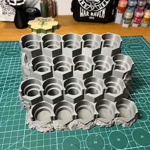 Puede incluir: Un soporte gris impreso en 3D para botes de pintura con m&uacute;ltiples ranuras hexagonales. El soporte est&aacute; dise&ntilde;ado para organizar y almacenar botes de pintura peque&ntilde;os, con una base texturizada y rocosa. El fondo incluye botes de pintura y una bolsa negra con el texto "WAR RAVEN FORGE".