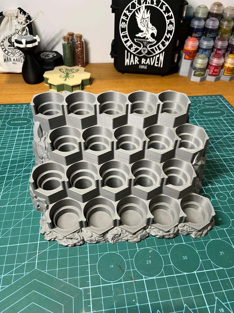 Puede incluir: Un soporte gris impreso en 3D para botes de pintura con m&uacute;ltiples ranuras hexagonales. El soporte est&aacute; dise&ntilde;ado para organizar y almacenar botes de pintura peque&ntilde;os, con una base texturizada y rocosa. El fondo incluye botes de pintura y una bolsa negra con el texto "WAR RAVEN FORGE".