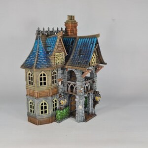 Op de afbeelding: Een gedetailleerd miniatuur fantasiehuis met een blauw pannendak, boogvensters en een stenen gevel. Het huis heeft meerdere niveaus, een schoorsteen en decoratieve elementen.