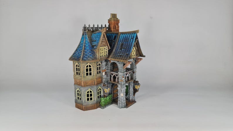 Op de afbeelding: Een gedetailleerd miniatuur fantasiehuis met een blauw pannendak, boogvensters en een stenen gevel. Het huis heeft meerdere niveaus, een schoorsteen en decoratieve elementen.