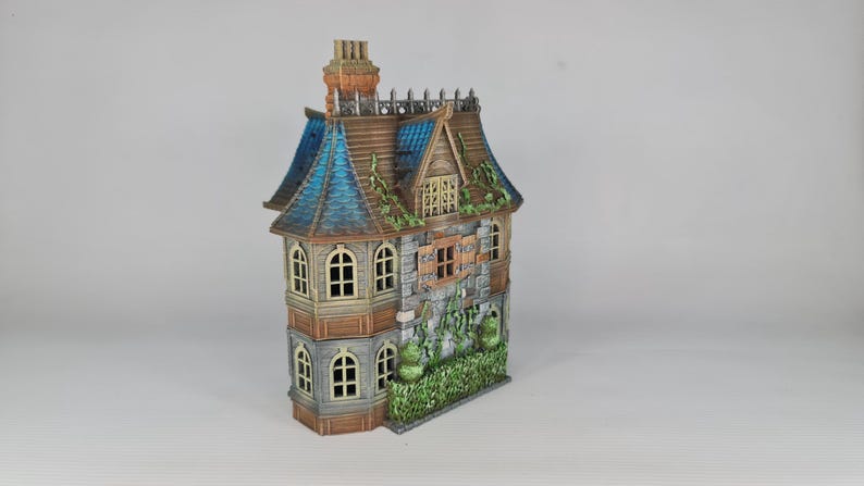 Op de afbeelding: Een gedetailleerd miniatuurhuis met een verweerde uitstraling. Het heeft een blauw en bruin dak, stenen muren en gewelfde ramen. Groen gebladerte accentueert de buitenkant en cre&euml;ert een gotische esthetiek. De schoorsteen is oranje.