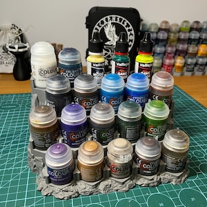 Puede incluir: Una colecci&oacute;n de botes de pintura Citadel Colour y Vallejo en varios colores, incluyendo azul, verde, marr&oacute;n y morado. Las pinturas est&aacute;n dispuestas sobre un expositor gris texturizado. En el fondo, una caja negra con un dise&ntilde;o blanco.