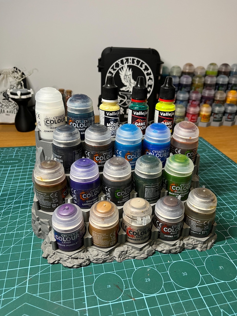 Puede incluir: Una colecci&oacute;n de botes de pintura Citadel Colour y Vallejo en varios colores, incluyendo azul, verde, marr&oacute;n y morado. Las pinturas est&aacute;n dispuestas sobre un expositor gris texturizado. En el fondo, una caja negra con un dise&ntilde;o blanco.