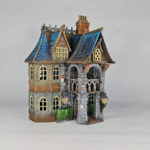 Op de afbeelding: Een gedetailleerd miniatuurhuis met een blauw pannendak, gewelfde ramen en een stenen gevel. Het gebouw met meerdere verdiepingen heeft decoratieve lantaarns en een schoorsteen. Het ontwerp suggereert een fantasie- of gotisch thema.