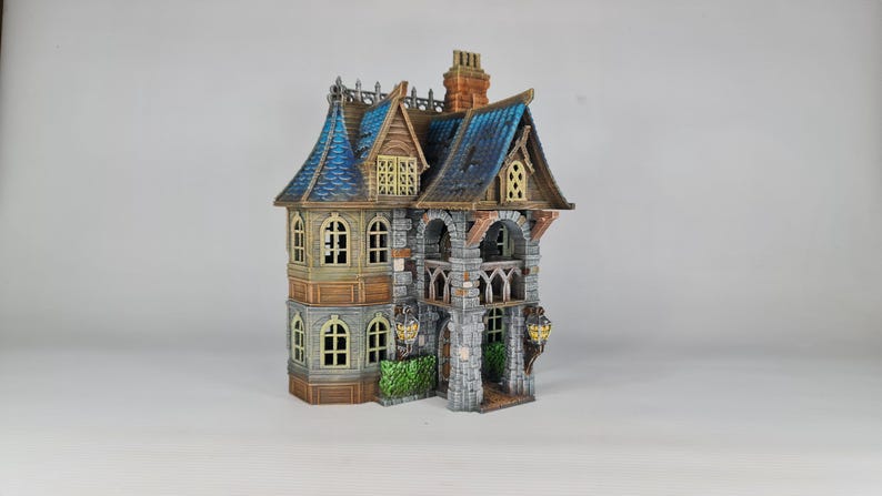 Op de afbeelding: Een gedetailleerd miniatuurhuis met een blauw pannendak, gewelfde ramen en een stenen gevel. Het gebouw met meerdere verdiepingen heeft decoratieve lantaarns en een schoorsteen. Het ontwerp suggereert een fantasie- of gotisch thema.