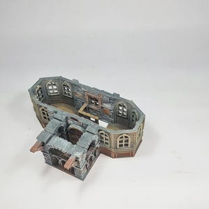 Op de afbeelding: Een miniatuurgebouwmodel met meerdere niveaus en een verweerde uitstraling. De structuur heeft boogvensters, een stenen gevel en een houten interieur. Het model is geschilderd in tinten grijs, bruin en beige, wat wijst op een middeleeuws of fantasiethema.