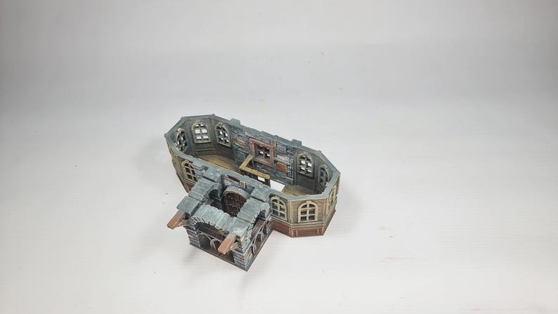 Op de afbeelding: Een miniatuurgebouwmodel met meerdere niveaus en een verweerde uitstraling. De structuur heeft boogvensters, een stenen gevel en een houten interieur. Het model is geschilderd in tinten grijs, bruin en beige, wat wijst op een middeleeuws of fantasiethema.