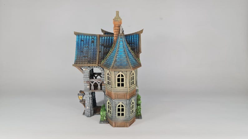 Op de afbeelding: Een miniatuurhuis met meerdere verdiepingen, een blauw pannendak en een toren. Het huis heeft boogvensters, een stenen balkon en een schoorsteen. De buitenkant is geschilderd in bruin-, beige- en grijstinten, met een lantaarn aan de zijkant.