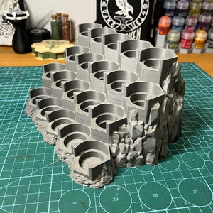 Puede incluir: Un soporte de pintura en miniatura gris impreso en 3D con m&uacute;ltiples ranuras hexagonales, dise&ntilde;ado para parecerse a un acantilado rocoso. El soporte est&aacute; dise&ntilde;ado para contener varios botes de pintura. El soporte est&aacute; sobre una esterilla de corte verde.