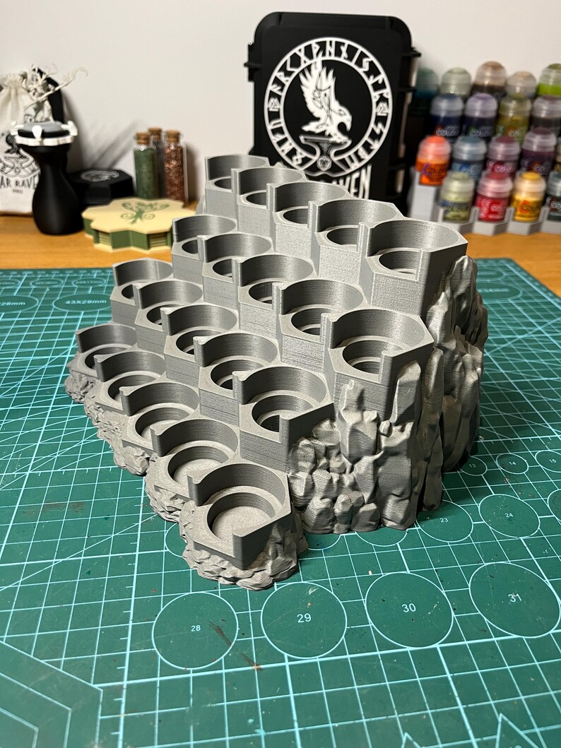Puede incluir: Un soporte de pintura en miniatura gris impreso en 3D con m&uacute;ltiples ranuras hexagonales, dise&ntilde;ado para parecerse a un acantilado rocoso. El soporte est&aacute; dise&ntilde;ado para contener varios botes de pintura. El soporte est&aacute; sobre una esterilla de corte verde.