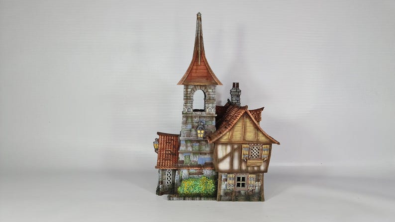 May include: A detailed miniature model of a medieval-style building with a tall steeple and a red tile roof. The building features stone and wood construction, with small windows and decorative details. The model includes a small garden.
