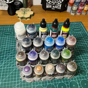 Puede incluir: Una colecci&oacute;n de botes de pintura en miniatura de varios colores, dispuestos en un organizador de pl&aacute;stico gris. Los botes est&aacute;n etiquetados con nombres de marcas y nombres de colores. Una bolsa negra con el texto "WAR RAVEN" est&aacute; en el fondo.