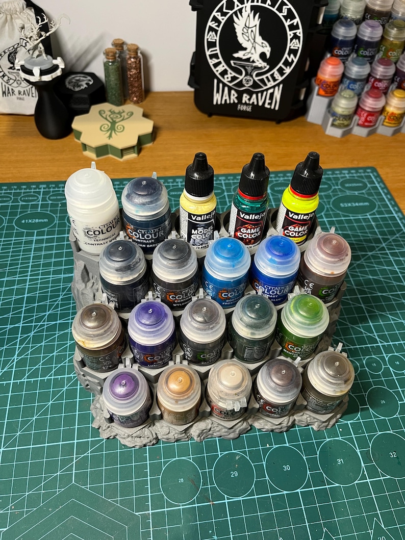 Puede incluir: Una colecci&oacute;n de botes de pintura en miniatura de varios colores, dispuestos en un organizador de pl&aacute;stico gris. Los botes est&aacute;n etiquetados con nombres de marcas y nombres de colores. Una bolsa negra con el texto "WAR RAVEN" est&aacute; en el fondo.