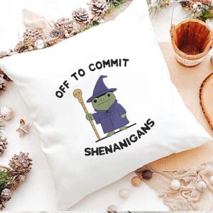K&ouml;nnte beinhalten: Wei&szlig;es quadratisches Kissen mit dem gestickten Text "OFF TO COMMIT SHENANIGANS". Das Kissen zeigt einen Cartoon-Frosch, der als Zauberer gekleidet ist, einen Stab h&auml;lt und einen lila Umhang und Hut tr&auml;gt.