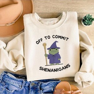 K&ouml;nnte beinhalten: Cremefarbenes Sweatshirt mit dem Text "OFF TO COMMIT SHENANIGANS" und einem Cartoon-Frosch, der als Zauberer verkleidet ist. Der Frosch tr&auml;gt einen lila Umhang und Hut und h&auml;lt einen Stab. Ein beiger Hut und blaue Jeans sind ebenfalls im Bild.