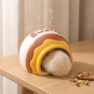 Può includere: Una casetta per criceti in ceramica a forma di ciambella. La casa è bianca, marrone e gialla, con la scritta "sugary" sopra. Un piccolo criceto soffice è all'interno. Un cucchiaio di legno con semi è accanto.